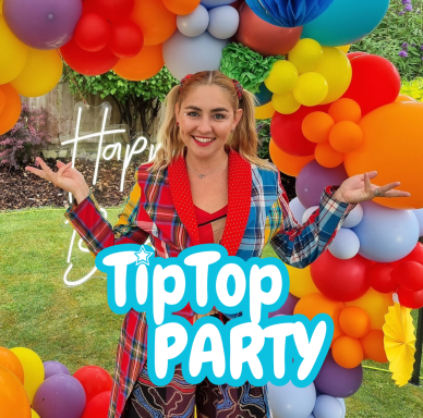 A smiling person in a multicoloured outfit stands under a vibrant balloon arch.